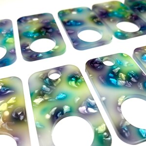 May include: A set of eight rectangular keychains with a hole at the top. Each keychain is made of clear resin with a swirling pattern of blue, green, and purple. The keychains have a round hole in the center.
