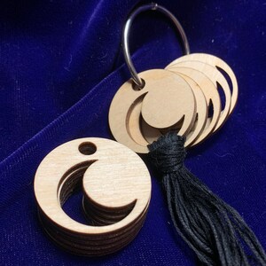 Może przedstawiać: Zestaw drewnianych zawieszek w kształcie księżyca z czarną frędzlą przymocowaną do srebrnego pierścienia. Zawieszki są warstwowe i mają wzór półksiężyca.