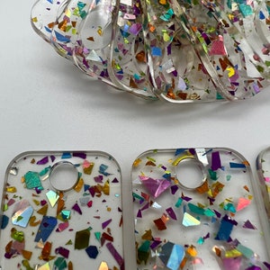 May include: A set of four clear resin keychains with a round hole and a confetti-like pattern of iridescent glitter in shades of purple, blue, green, gold, and orange.
