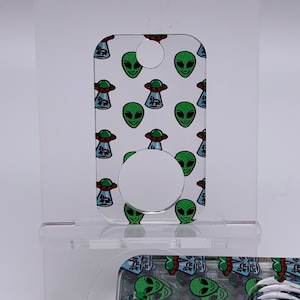 May include: A clear acrylic phone stand with a green alien and UFO pattern. The stand has a circular cutout for holding a phone.