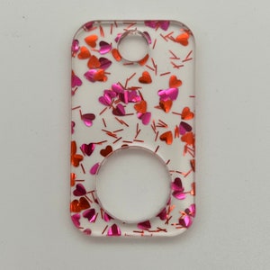 Op de afbeelding: Een transparante rechthoekige sleutelhanger met een glitterhartontwerp in roze en rood. De sleutelhanger heeft een gat aan de bovenkant om aan een sleutelring te bevestigen en een rond gat in het midden.