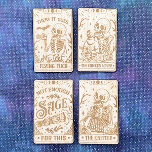 Op de afbeelding: Vier houten kaarten met illustraties van skeletten en tekst. De kaarten lezen "There it goes. My last flying fuck", "The Chicken Lover", "Not enough sage for this", en "The Knitter".