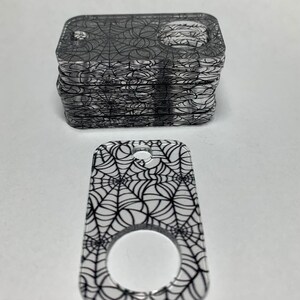 May include: A stack of clear acrylic keychains with a black spiderweb design. The keychain has a ring for attaching keys.