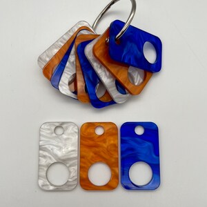 May include: A set of six colorful, square-shaped keychains with a hole in the center. The keychains are made of a marbled plastic material in white, blue, and orange.