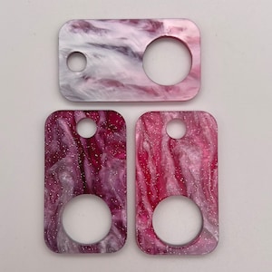 May include: Three pink and white marbled keychains with a hole at the top and a round cutout in the middle. The keychains have a glittery finish.