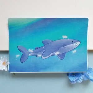 May include: A blue cartoon shark swims in a turquoise and blue ocean with white bubbles. The shark has a white belly and a white stripe on its back.