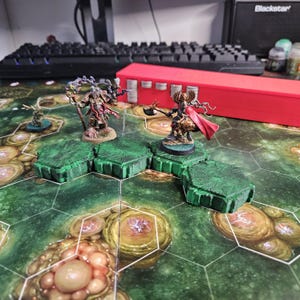 May include: A tabletop game scene featuring miniature figures on a hexagonal board. The figures include a warrior with a red cape and axe, and a wizard with a staff. The board has green terrain tiles and a detailed, otherworldly design.