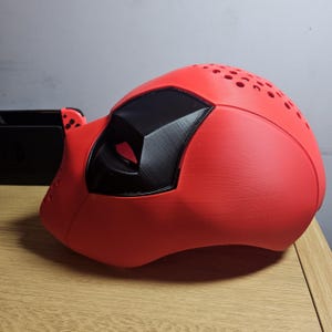 May include: A red and black mask with a smooth finish. The mask has a rounded shape with a black eye covering and small ventilation holes on top. The mask is likely a costume or cosplay accessory.