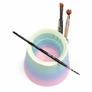 May include: A pastel-colored paint brush holder with a gradient of pink, blue, and green. Three paint brushes are in the holder, and one black paint brush is resting on the side. The holder is designed to keep brushes organized.