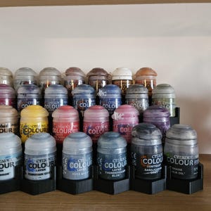 Peut inclure: Plusieurs pots de peinture Citadel Colour de différentes couleurs, dont du jaune, du rouge, du bleu et du gris. Les pots sont disposés sur des présentoirs noirs à plusieurs niveaux, avec des étiquettes visibles. L'image présente différents types de peinture.