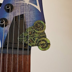 May include: Close-up of a guitar headstock with strings and tuning pegs. A green and gold-coloured, ornate, three-lobed design is affixed to the headstock. The headstock has a blue and white gradient design.
