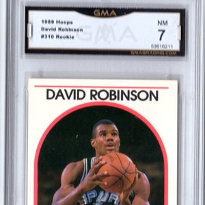 May include: A 1989 NBA Hoops trading card featuring David Robinson, a basketball player for the San Antonio Spurs. The card is in near mint condition and has a red, white, and black color scheme. The card features a photo of Robinson in his Spurs uniform, holding a basketball. The card also includes the text "David Robinson", "NBA Hoops", "Spurs", and "Center".