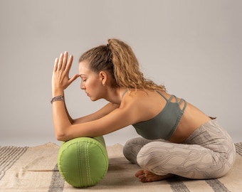 Op de afbeelding: Een vrouw in een grijze sportbeha en een witte, gepatroneerde legging zit op een groene yogabolste. Ze houdt haar handen voor haar gezicht bij elkaar, met haar ogen dicht.