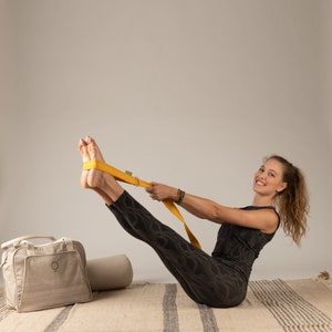 May include: A woman in black leggings and a black tank top is doing a yoga pose with a yellow yoga strap. She is smiling and looking at the camera. The yoga strap is wrapped around her feet and she is holding the ends of the strap in her hands.