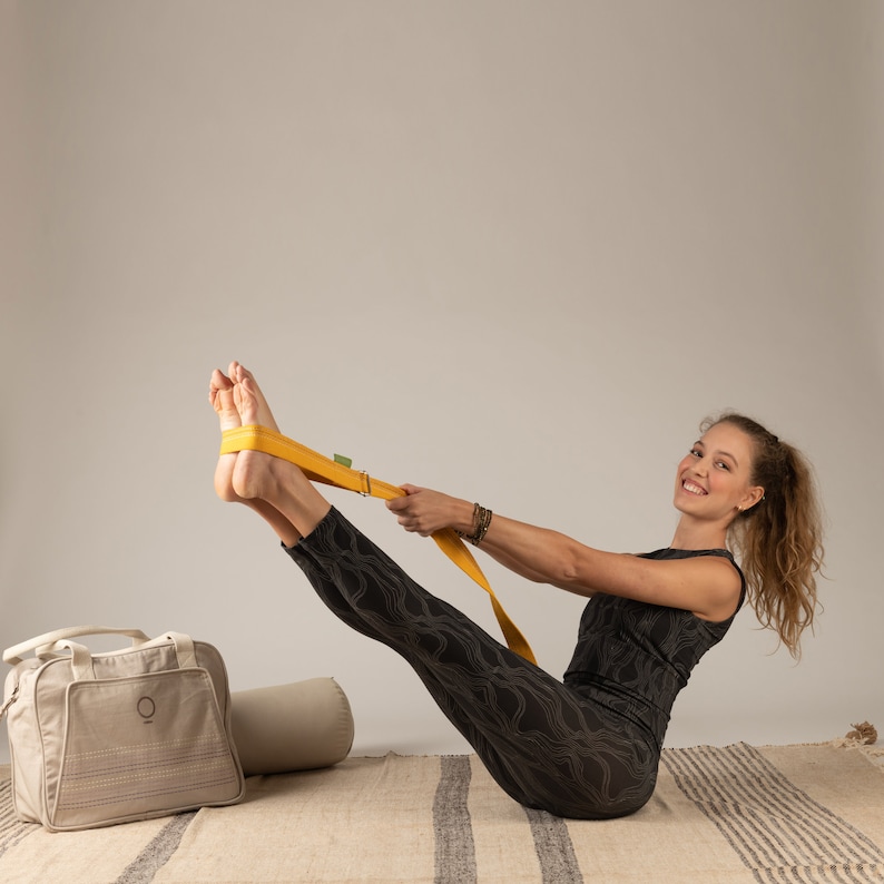 May include: A woman in black leggings and a black tank top is doing a yoga pose with a yellow yoga strap. She is smiling and looking at the camera. The yoga strap is wrapped around her feet and she is holding the ends of the strap in her hands.