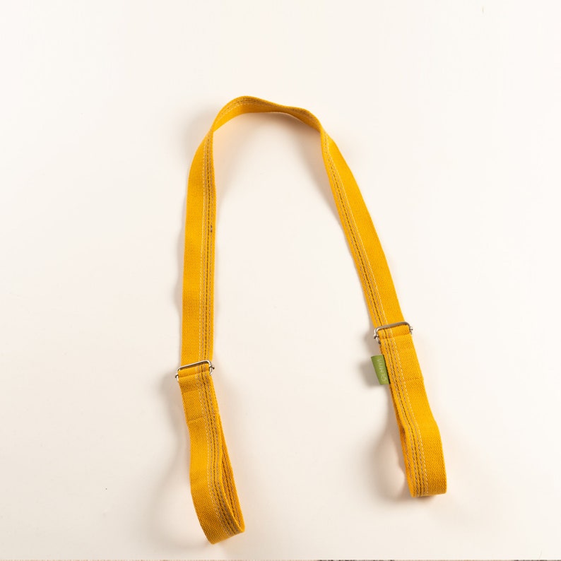 May include: A yellow fabric strap with two metal buckles. The strap is designed for carrying a bag or other object.