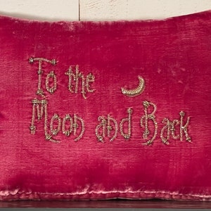 May include: A pink velvet pillow with a beaded inscription that reads "To the Moon and Back" with a crescent moon design.