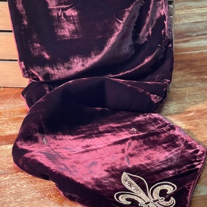 May include: A burgundy velvet throw pillow with a gold embroidered fleur-de-lis design. The pillow has a tassel on one corner.