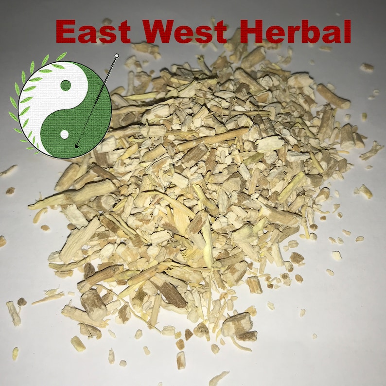 Op de afbeelding: Een close-up van een hoop gedroogde, lichtbruine en beige kruidenwortels. De wortels zijn gebroken in kleine stukjes en zijn verspreid over een wit oppervlak. De tekst "East West Herbal" staat in rode letters bovenaan de afbeelding.
