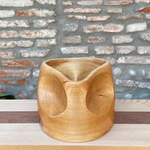 Può includere: Una ciotola di legno intagliata a mano con finitura marrone chiaro. La ciotola ha una forma unica e arrotondata con tre grandi ritagli circolari sui lati. L'interno della ciotola è liscio e curvo, e la venatura del legno è visibile.