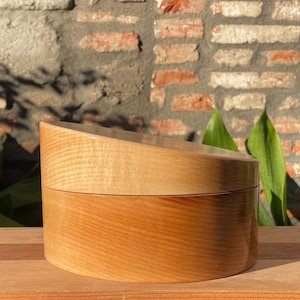 May include: A two-piece wooden box with a rounded lid. The box is made of light wood with a natural finish. The lid is slightly angled and has a smooth, polished surface.