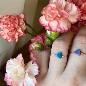 Puede incluir: Dos anillos delicados con piedras en forma de corazón. Las piedras son iridiscentes y tienen un tono azul y morado. Los anillos están hechos de alambre fino y se llevan en un dedo.