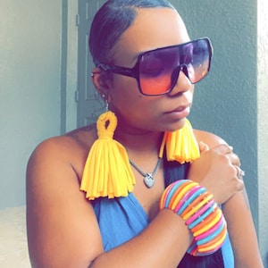 Könnte beinhalten: Eine Frau trägt eine große, schwarze, quadratische Sonnenbrille mit Verlaufsglas. Sie trägt ein blaues, ärmelloses Oberteil und hat gelbe Quastenohrringe und ein buntes, mehrlagiges Armkettchen am Handgelenk.