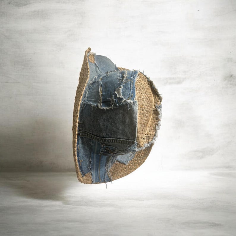 Puede incluir: Sombrero de paja con parches de mezclilla. El sombrero es de color beige natural con secciones de mezclilla azul cosidas en el ala y la copa. La mezclilla tiene un aspecto desgastado.
