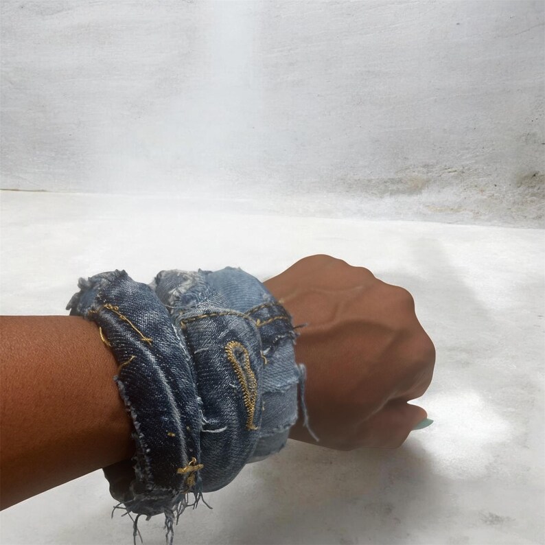 Puede incluir: Una pulsera de mezclilla hecha de jeans reciclados. La pulsera es de color azul claro con bordes deshilachados y costuras amarillas. La pulsera est&aacute; envuelta alrededor de una mu&ntilde;eca.