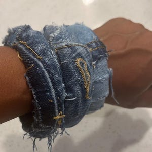 Puede incluir: Una pulsera de mezclilla hecha de jeans reciclados. La pulsera es de color azul oscuro con bordes deshilachados y costuras visibles. La pulsera est&aacute; envuelta alrededor de una mu&ntilde;eca, mostrando su textura y dise&ntilde;o. El estilo es casual y hecho a mano.