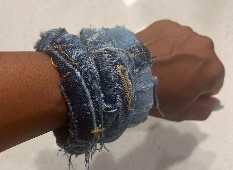 Puede incluir: Una pulsera de mezclilla hecha de jeans reciclados. La pulsera es de color azul oscuro con bordes deshilachados y costuras visibles. La pulsera est&aacute; envuelta alrededor de una mu&ntilde;eca, mostrando su textura y dise&ntilde;o. El estilo es casual y hecho a mano.