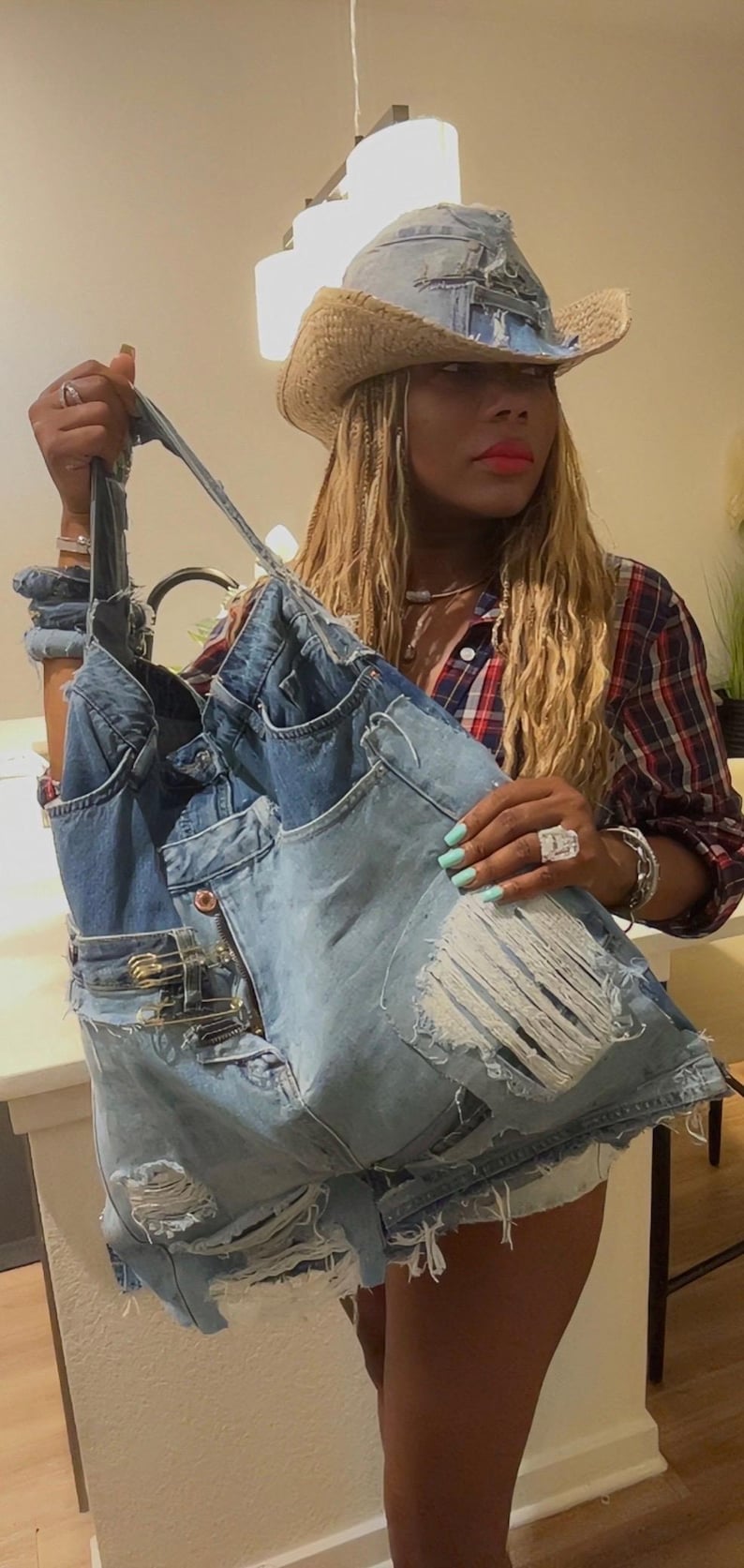 Puede incluir: Un bolso tote de mezclilla hecho de jeans reciclados, con bolsillos, detalles desgastados y cierre de cremallera. El bolso es sostenido por una persona. El bolso se combina con un sombrero de vaquero de mezclilla a juego.