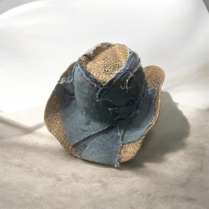 Puede incluir: Un sombrero de vaquero de paja con un dise&ntilde;o de patchwork de mezclilla. El sombrero presenta un color paja natural con parches de mezclilla azul claro cosidos en la corona y el ala. La mezclilla tiene un borde deshilachado y desgastado.
