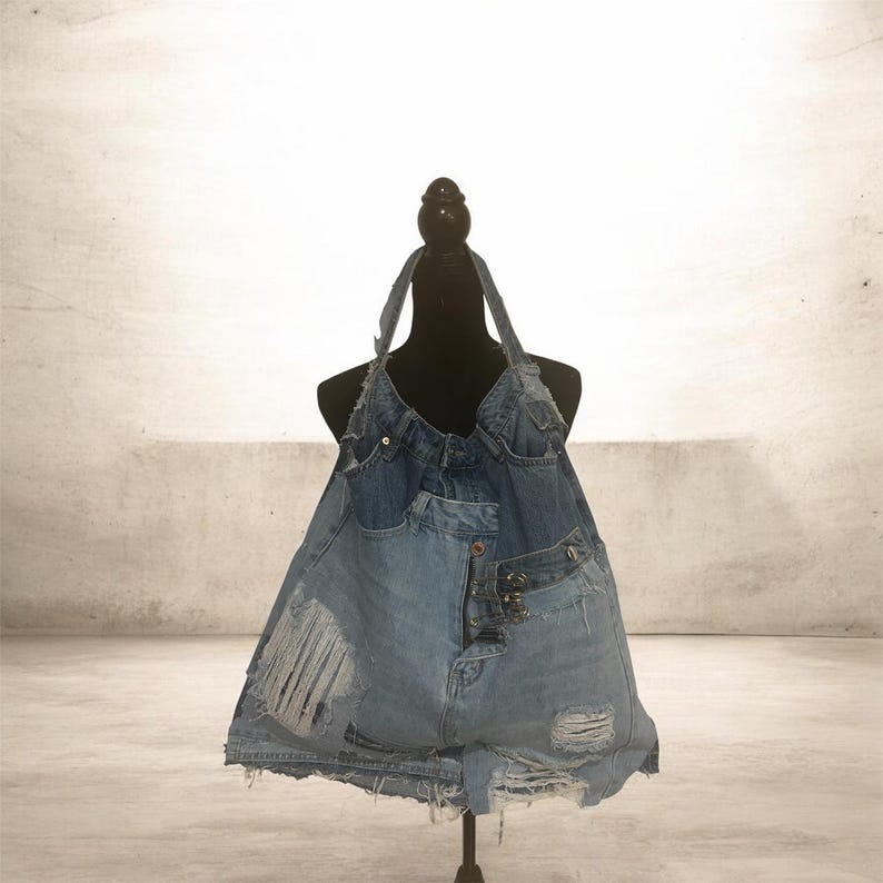 Puede incluir: Un bolso tote de mezclilla hecho a mano, construido con jeans azules reciclados. El bolso presenta una correa estilo halter y un dise&ntilde;o de patchwork con detalles desgastados, incluyendo secciones rasgadas y bolsillos visibles. El bolso se exhibe en un maniqu&iacute; negro.