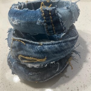 Puede incluir: Tres pulseras de mezclilla apiladas. Cada pulsera est&aacute; hecha de tela de mezclilla azul con bordes deshilachados y costuras amarillas. Las pulseras est&aacute;n apiladas, creando un efecto en capas.