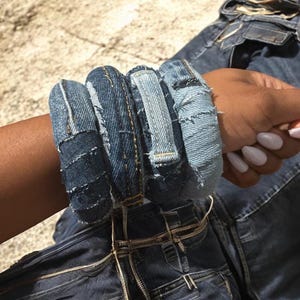 May include: A set of four denim bracelets, each crafted from repurposed blue jean fabric. The bracelets feature a patchwork design with visible stitching and frayed edges, showcasing a unique, handcrafted aesthetic. The bracelets are stacked on a wrist.