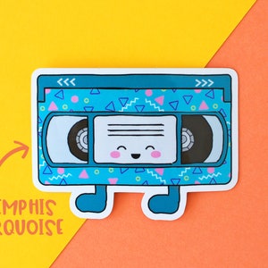 May include: A blue and turquoise VHS tape sticker with a happy face and a geometric pattern. The sticker says "<< >>" on the top and "Memphis Turquoise" on the bottom.