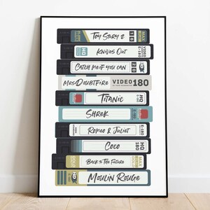 Może przedstawiać: Oprawiony plakat przedstawiający stos kaset VHS w stylu retro. Każda kaseta jest oznaczona tytułem filmu, np. "Toy Story 2" i "Romeo & Juliet". Kasety są w r&oacute;żnych kolorach, z białym tekstem na czarnym tle.