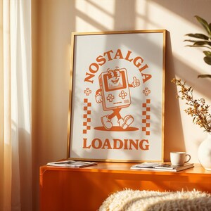 May include: A framed poster with a retro-style illustration of a walking video game console with the words "NOSTALGIA LOADING" in orange. The poster is in a gold frame and is displayed on an orange cabinet. A white vase with greenery and a coffee cup are also on the cabinet.