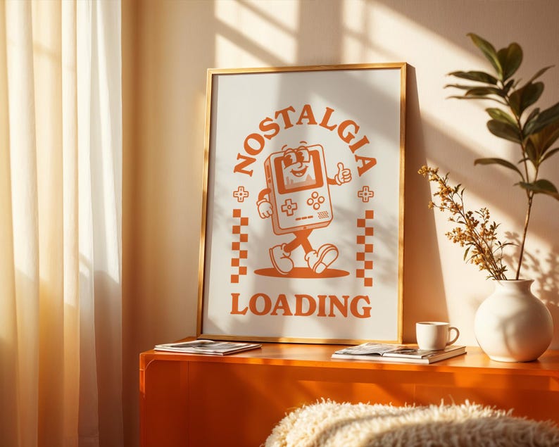 May include: A framed poster with a retro-style illustration of a walking video game console with the words "NOSTALGIA LOADING" in orange. The poster is in a gold frame and is displayed on an orange cabinet. A white vase with greenery and a coffee cup are also on the cabinet.