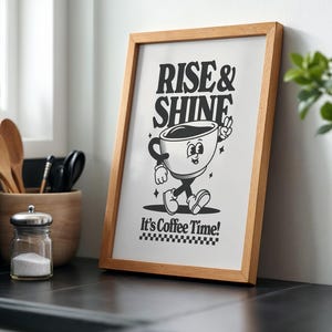 May include: A framed print with a black and white illustration of a coffee cup with legs and arms. The text "Rise & Shine" is above the cup and "It's Coffee Time!" is below the cup.