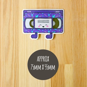 May include: A purple and blue VHS tape sticker with a smiling face and the text "<<< >>>" on the top. The sticker is approximately 75mm x 55mm.