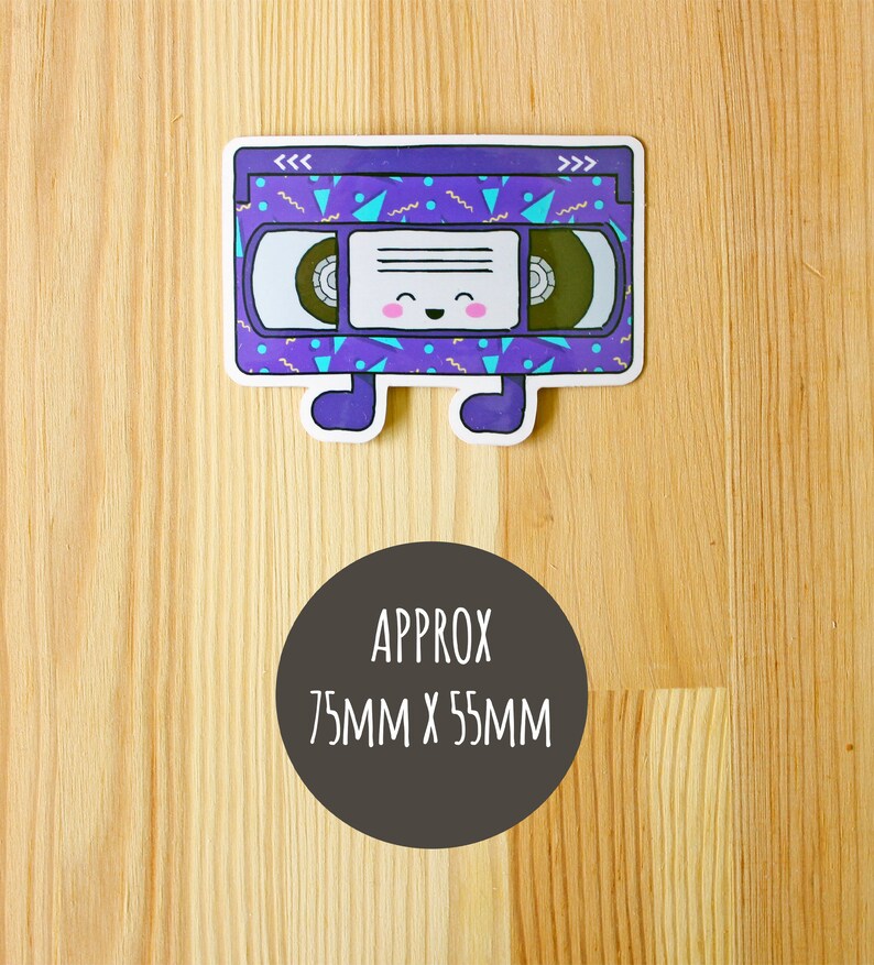 May include: A purple and blue VHS tape sticker with a smiling face and the text "<<< >>>" on the top. The sticker is approximately 75mm x 55mm.