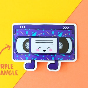 May include: A purple and blue VHS tape with a happy face and a 90s style geometric pattern. The tape has the words "<<<" and ">>>" on the top.