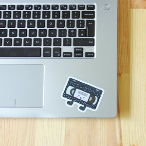 May include: A black and white cassette tape sticker with a cute cartoon face and pink cheeks. The sticker is on a silver laptop computer.
