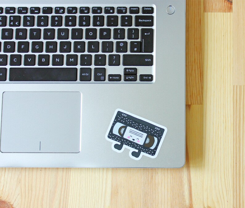 May include: A black and white cassette tape sticker with a cute cartoon face and pink cheeks. The sticker is on a silver laptop computer.