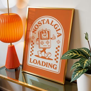 May include: Orange framed print with the words "Nostalgia Loading" and a cartoon game console character. The print is displayed on a glass-topped table with an orange lamp and a potted plant.