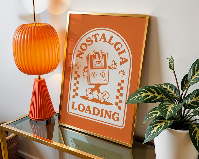 May include: Orange framed print with the words "Nostalgia Loading" and a cartoon game console character. The print is displayed on a glass-topped table with an orange lamp and a potted plant.