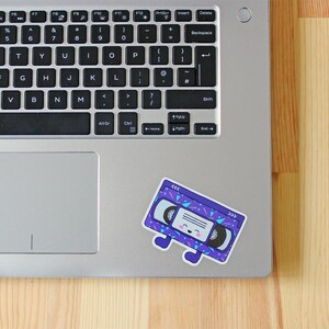 May include: A purple and blue cassette tape sticker with a smiling face and a geometric pattern. The sticker is on a silver laptop.