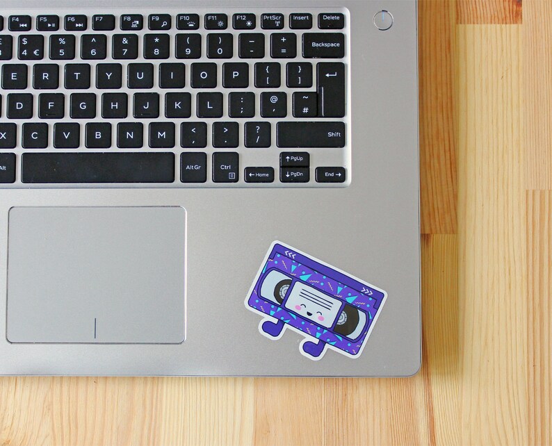 May include: A purple and blue cassette tape sticker with a smiling face and a geometric pattern. The sticker is on a silver laptop.